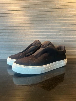 Axel Arigato skor - Stilrena svarta slip-on sneakers i mjuk mocka med tjock vit sula. Skorna har minimalistisk design utan synliga snören och en låg silhuett som ger en clean look. Perfekta för dig som gillar enkel och modern stil.