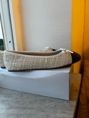 Beige ballerinaskor med lackad tå - Snygga ballerinaskor i beige tyg med struktur och rundad, svart lackad tå. Dekorativa små rosetter framtill ger en söt touch. Skorna har låg profil och klassisk form, perfekta för en trendig och stilren look.