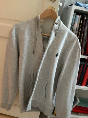 Grå hoodie med dragkedja från Polo - Snygg ljusgrå hoodie med dragkedja från Polo Ralph Lauren. Klassisk design med huva och dragsnören, två fickor framtill och den ikoniska svarta Polo-loggan broderad på bröstet. Perfekt för en avslappnad och stilren look.