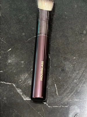 Hourglass sminkborste i metallic - Snygg sminkborste från Hourglass med ett elegant metalliclila handtag och täta, mjuka borststrån i vitt och beige. Perfekt för applicering av puder, rouge eller highlighter. Handtaget har en lyxig känsla och Hourglass-loggan i guld.