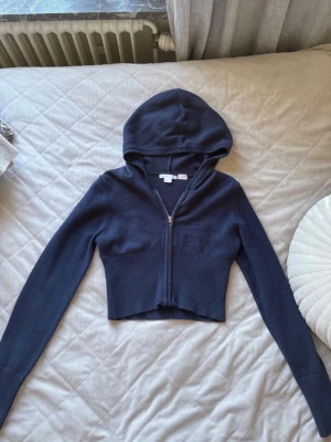 Mörkblå croppad hoodie med dragkedja - Snygg mörkblå croppad hoodie med dragkedja framtill och långa ärmar. Modellen har ribbad midja och är tillverkad i ett mjukt stickat material. Perfekt för en avslappnad och trendig look.