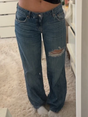 Blå raka jeans med hål - Säljer ett par blå raka jeans med ett stort slitet hål på höger lår. Jeansen har klassisk femficksdesign, hög midja och knappgylf. Perfekta för en avslappnad och trendig look.