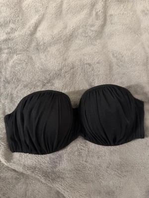 Svart axelbandslös bandeau-bh - Säljer en svart axelbandslös bandeau-bh med lätt rynkad framsida och vadderade kupor. Bh:n har breda sidor för extra stöd och stängs med hyskor i ryggen. Materialet är mjukt och stretchigt, perfekt under toppar utan axelband.