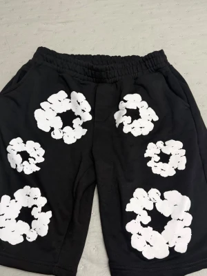 Denim tears shorts  storlek S - Storlek M Svarta shorts med elastisk midja och stora vita blommönster tryckta över hela plagget. Shortsen har fickor på sidorna och en bakficka, perfekt för en avslappnad och trendig look.