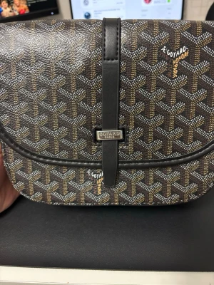 Brun handväska med monogram - Snygg brun handväska med klassiskt monogrammönster i vitt och gult. Väskan har rundad form, lock och svart läderrem med metallspänne. Perfekt för dig som gillar exklusiva accessoarer och vill sticka ut.
