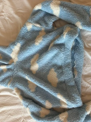 Blå fluffig tröja med molnmönster - Supermjuk och fluffig stickad tröja från Lindex i ljusblått med vita moln. Tröjan har lång ärm och avslappnad passform, perfekt för att mysa i. Materialet är teddy/fleece-liknande och ger en riktigt cozy känsla.