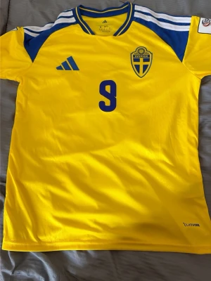 Svensk landslagströja Adidas gul - Gul svensk landslagströja från Adidas med blå detaljer och nummer 9 på bröstet. Tröjan har blått landslagsemblem, vita och blå ränder på axlarna samt ClimaCool-material för bra ventilation. Perfekt för fotboll eller att bära på matchdag.