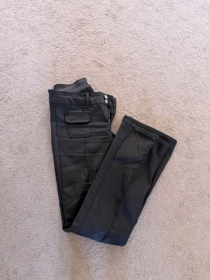 Svarta coated jeans med fickdetaljer - Snygga svarta jeans med coated finish och raka ben. Framtill finns två fickor med lock och knapp, samt klassisk knappgylf. Materialet ger en lätt glansig look som sticker ut. Perfekta för dig som gillar en trendig och edgy stil.
