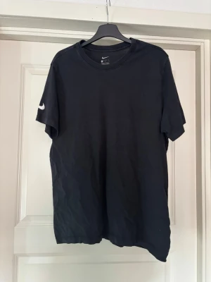 Svart Nike t-shirt med logga - En klassisk svart t-shirt från Nike med vit swoosh-logga på ärmen. T-shirten har rund halsringning och är tillverkad i mjuk bomull för en bekväm känsla. Enkel och stilren design som passar till det mesta.