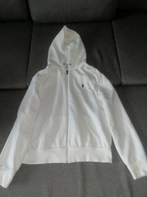 Vit hoodie med dragkedja från Polo Ralph Lauren - Snygg vit hoodie med dragkedja från Polo Ralph Lauren. Klassisk design med huva, fickor framtill och den ikoniska lilla loggan broderad på bröstet. Perfekt för en clean och stilren look. Tillverkad i mjukt bomullsmaterial.