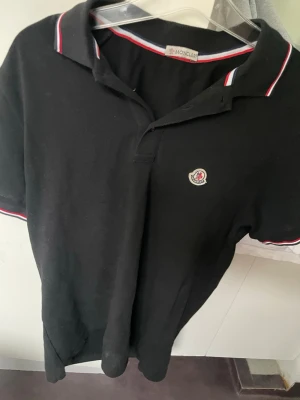 Svart Moncler pikétröja med röd/vita detaljer - Klassisk svart pikétröja från Moncler med korta ärmar och krage. Tröjan har röda och vita ränder på krage och ärmslut samt Moncler-logga på bröstet. Perfekt för en stilren och sportig look. Loggan ör Scan bar