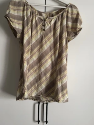 Randig beige och brun blus - Luftig blus med diagonala ränder i beige, brunt och ljusgult. Modellen har korta ärmar, rund halsringning med liten knappdetalj framtill och ett avslappnat fall. Perfekt för varma dagar och enkel att styla till jeans eller kjol.