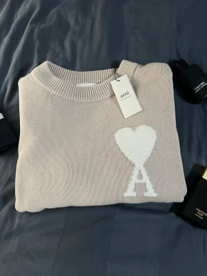 Beige stickad tröja från Ami - Snygg beige stickad tröja från Ami med rund halsringning och ribbade muddar. Tröjan har ett vitt hjärta och bokstaven 'A' broderat på bröstet. Perfekt för dig som gillar stilrena och trendiga plagg.