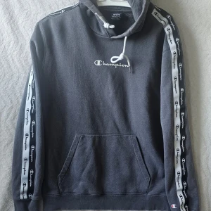 Grå Champion hoodie med logga - Snygg grå hoodie från Champion med vit broderad logga på bröstet och vita band med Champion-tryck längs ärmarna. Klassisk känguruficka framtill och justerbar huva med dragsko. Perfekt för en avslappnad streetwear-look.