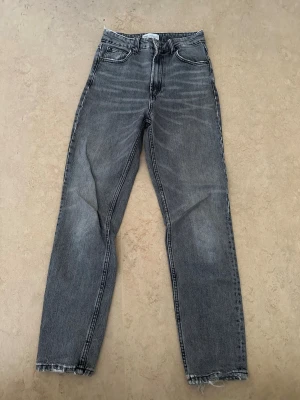 Grå raka jeans från ZARA - Säljer ett par grå raka jeans från ZARA med hög midja och klassisk femficksmodell. Jeansen har slitna detaljer vid bensluten och är tillverkade i ett mjukt denimtyg. Perfekta för en avslappnad och trendig look.
