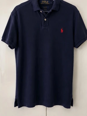 Marinblå pikétröja från Polo Ralph Lauren - Klassisk marinblå pikétröja från Polo Ralph Lauren med röd broderad logga på bröstet. Tröjan har korta ärmar, krage och två knappar framtill. Tillverkad i mjuk bomull för en bekväm och snygg look.