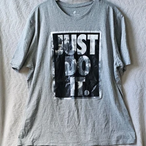 Grå Nike t-shirt med tryck XXL - Säljer en grå t-shirt från Nike i storlek XXL med stort svartvitt tryck på bröstet där det står 'JUST DO IT.'. Klassisk rund hals och korta ärmar. Perfekt för dig som gillar sportig och avslappnad stil.