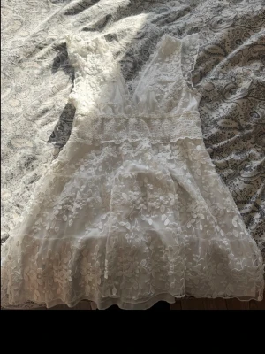 Vit kort spetsklänning med blommor - Superfin vit kort klänning med v-ringning och bred spets i midjan. Klänningen har blommiga spetsdetaljer över hela tyget och är ärmlös. Perfekt för sommarens alla tillfällen och ger en romantisk vibe.