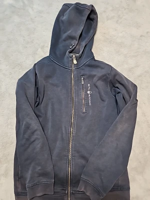 Mörkblå hoodie med dragkedja - Snygg mörkblå hoodie med dragkedja och huva. Framtill finns en vertikal bröstficka med dragkedja och tryckt text 'Sail Racing'. Perfekt för en avslappnad och sportig stil. Mjukt material och ribbade muddar vid ärmslut och nederkant.