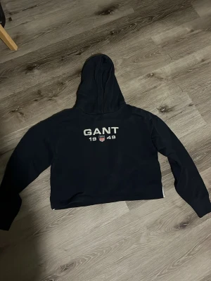 Svart hoodie från GANT med tryck - Snygg svart hoodie från GANT med stort vitt tryck på bröstet där det står 'GANT 1949' och en liten sköld i mitten. Hoodien har huva och långa ärmar. Perfekt för en avslappnad och sportig stil. Magtröja