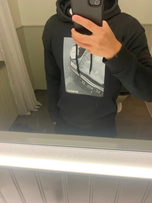 Svart hoodie med biltryck - Säljer en svart hoodie med stor grafisk print av en klassisk bil på bröstet. Hoodien har huva med dragsko och ribbade muddar vid ärmar och midja. Perfekt för dig som gillar streetwear och coola motiv.