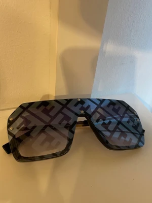 F solglasögon med mönster - Stora, svarta solglasögon med rektangulär form och tydligt mönster på glaset. Glasögonen har en modern design och ger en cool statement-look. Perfekta för dig som vill sticka ut med accessoarerna.