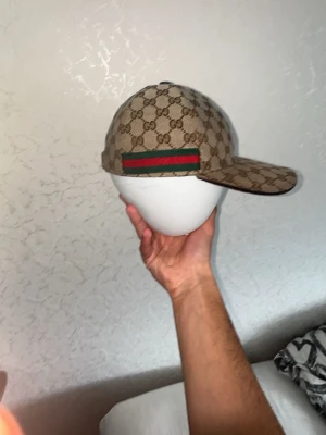 Äkta Beige Gucci Keps  - Säljer en beige keps från Gucci med det klassiska GG-monogrammönstret över hela kepsen. På sidan finns en grön och röd rand som signaturdetalj. Kepsen har böjd skärm och är tillverkad i canvasmaterial. Storlek M.