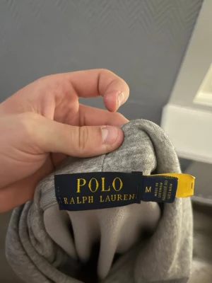 Grå hoodie från Polo Ralph Lauren - Snygg grå hoodie från Polo Ralph Lauren i storlek M. Klassisk design med huva, magficka och diskret broderad logga på bröstet. Tillverkad i mjukt bomullsmaterial som är skönt att ha på sig. Perfekt för en avslappnad och stilren look.