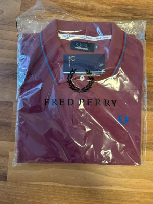 Vinröd piké från Fred Perry medium - Säljer en vinröd pikétröja från Fred Perry med blå och röda detaljer på kragen samt den klassiska loggan broderad på bröstet. Tröjan har korta ärmar och är tillverkad i bomull för en skön och stilren look.