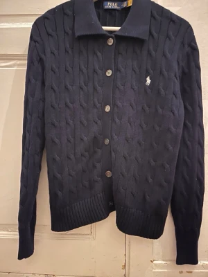 Mörkblå kabelstickad kofta Polo Ralph Lauren - Kabelstickad kofta i mörkblått från Polo Ralph Lauren med klassisk krage och knäppning framtill. Liten broderad logga på bröstet och ribbade muddar vid ärmslut och nederkant. Perfekt för en stilren och tidlös look.