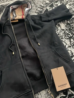 Svart zip hoodie från Burberry - Snygg svart hoodie från Burberry med dragkedja och klassisk huva. Insidan av huvan har Burberrys ikoniska rutmönster i beige, rött och vitt. Hoodien har fickor framtill och guldfärgade detaljer på snörena. Perfekt för en stilren och lyxig look.