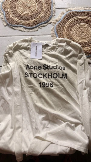 Vit långärmad tröja Acne Studios - Säljer en vit långärmad tröja från Acne Studios med trycket 'STOCKHOLM 1996' i svart på bröstet. Tröjan har en klassisk passform och är gjord i mjukt bomullsmaterial. Perfekt för dig som gillar minimalistisk och stilren design.