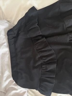 Svart kjol med volangdetalj - Snygg svart kortkjol med bred volangdetalj framtill. Kjolen har en enkel design och är perfekt för att skapa en trendig look. Tillverkad i ett mjukt tyg som ger en bekväm passform.