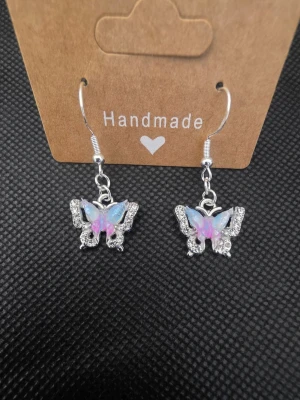 Silvriga fjärilsörhängen med glitter - Säljer ett par handgjorda örhängen i form av fjärilar. Örhängena är silvriga med gnistrande stenar och har vingar i skimrande blått och rosa. De har krokfäste och är perfekta för dig som gillar söta och unika accessoarer.