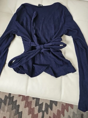 Mörkblå långärmadtopp med knytband - Snygg mörkblå tröja med långa ärmar och knytband i midjan. Toppen har v-ringning och är tillverkad i ett mjukt, lätt strukturerat material. Perfekt att styla med jeans eller kjol för en trendig look.