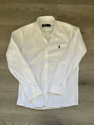 Vit skjorta från Polo Ralph Lauren - Klassisk vit skjorta från Polo Ralph Lauren med broderad logga på bröstet. Skjortan har lång ärm, knappar framtill och en stilren krage. Perfekt för en clean och tidlös look.