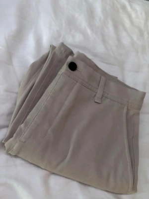 Beigea långbyxor med fickor - Stilrena beige kostymbyxor med klassisk passform och praktiska fickor framtill. De har bälteshällor och stängs med en svart knapp. Perfekta för varma dagar och enkla att matcha med olika outfits.