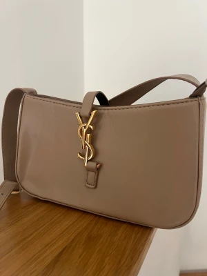 Beige axelremsväska från Saint Laurent - Snygg beige axelremsväska från Saint Laurent i slätt läder med guldigt YSL-emblem framtill. Väskan har en stilren, rektangulär form och justerbar axelrem. Perfekt för dig som gillar klassisk och trendig design.