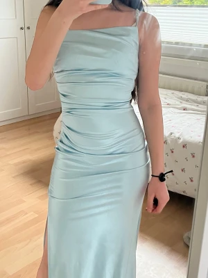 Ljusblå långklänning i satin - Supersnygg ljusblå långklänning i satin med smala axelband och slits på sidan. Klänningen har en figurnära passform och en elegant, glansig yta som ger en lyxig känsla. Perfekt för speciella tillfällen när du vill sticka ut.