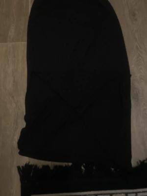 Svart kjol med öppning - Svart tygstycke i rektangulär form med dekorativa fransar längs ena kanten. Perfekt som accessoar till olika outfits eller som dekoration. Materialet ser ut att vara mjukt tyg.