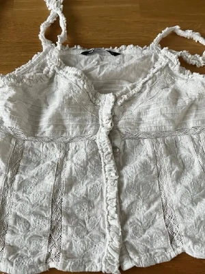 Vit spetsdetaljerad linnetopp från Zara - Supersöt vit linnetopp från Zara med spetsdetaljer, volanger och knappar framtill. Axelbanden är tvinnade och toppen har ett romantiskt broderat mönster. Perfekt för varma dagar och ger en bohemisk vibe.