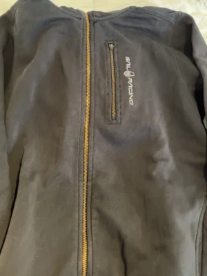 Svart hoodie från Sail Racing - Svart hoodie från Sail Racing med dragkedja framtill och en bröstficka med dragkedja. Loggan är tryckt i vitt på bröstet. Hoodien har en enkel och sportig design, perfekt för en avslappnad stil. Materialet ser ut att vara mjukt och bekvämt. Kontakta för intresse/frågor!
