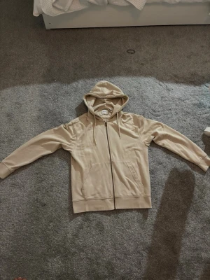 Beige hoodie med dragkedja - Snygg beige hoodie från Jack & Jones med dragkedja framtill och två fickor. Klassisk modell med huva och justerbara snören. Tillverkad i mjukt bomullsmaterial som är skönt att ha på sig. Perfekt för en avslappnad och stilren look.