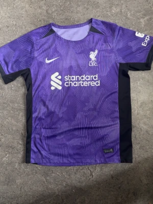 Lila Liverpool matchtröja Nike - Snygg lila Liverpool FC matchtröja från Nike med vita detaljer och klubbmärke broderat på bröstet. Tröjan har svart panel på sidorna, korta ärmar och tryckt sponsorlogga framtill. Perfekt för fotbollsträning eller att bära till vardags.