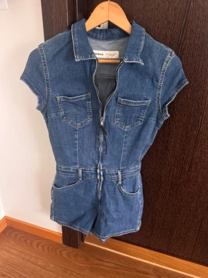 Kortärmad jeansjumpsuit med dragkedja - Snygg blå jeansjumpsuit i denim med korta ärmar och shortsben. Dragkedja framtill, fyra fickor fram och två bak. 