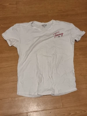 Vit Tommy Hilfiger t-shirt med tryck - INSTABOX!!! Vit t-shirt från Tommy Hilfiger med färgglatt tryck på ryggen och rosa logga framtill. Klassisk passform och rund hals. Perfekt för dig som gillar streetstyle och vill ha en enkel men snygg tee med coola detaljer.