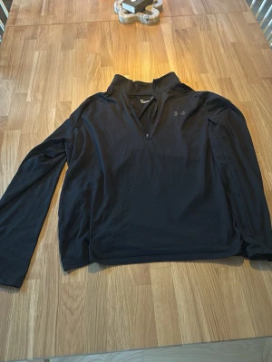 Svart långärmad tröja med halv dragkedja - Svart långärmad tröja från Under Armour med halv dragkedja framtill och hög krage. Tillverkad i ett mjukt och stretchigt material som passar perfekt för träning eller chill. Diskret logga på bröstet och enkel, stilren design.