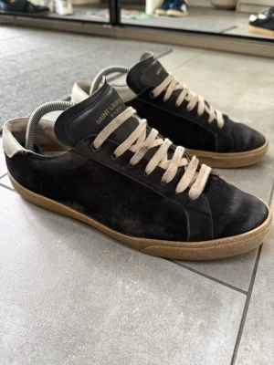 Svarta Saint Laurent sneakers - Svarta sneakers från Saint Laurent med vit häl och beige snörning. Skorna har en klassisk låg profil, ovandel i mocka och detaljer i läder. Yttersulan är i gummi med en vintage-look. Perfekta för en stilren och trendig vardagsstil. Skorna är slitna undertill men hela på insidan. Välanvändna men mycket kvar att ge.