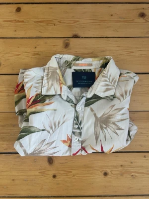 Blommig kortärmad skjorta Scotch & Soda - Säljer en beige kortärmad skjorta från Scotch & Soda med tropiskt blommönster i grönt, orange och rött. Skjortan har klassisk krage och knäppning framtill. Perfekt för sommardagar och ger en avslappnad vibe.