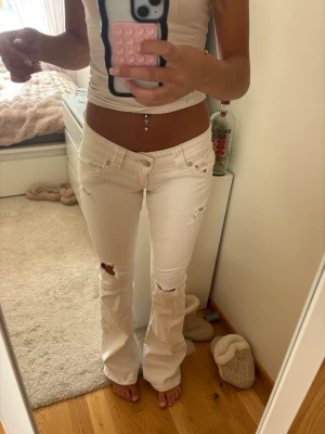 Vita bootcut jeans med slitningar - Säljer ett par vita bootcut jeans med slitningar på låren och knäna. Jeansen har låg midja, fem fickor och råa detaljer för en trendig look. Lite smuts på ena benet men inget som man lägger märke till.